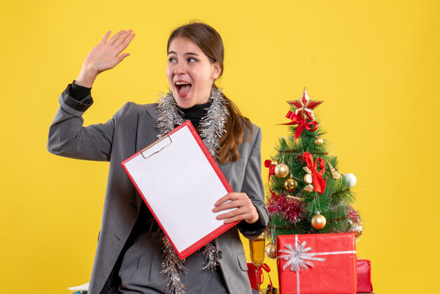 知识分子前视图微笑的女孩与文件招呼某人站在圣诞树和礼物鸡尾酒欢呼文件微笑