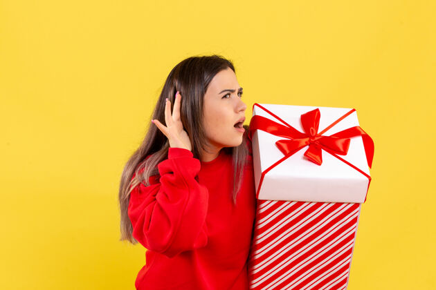 听年轻女子拿着圣诞礼物在盒子里 在黄色的墙上聆听的正视图漂亮快乐节日