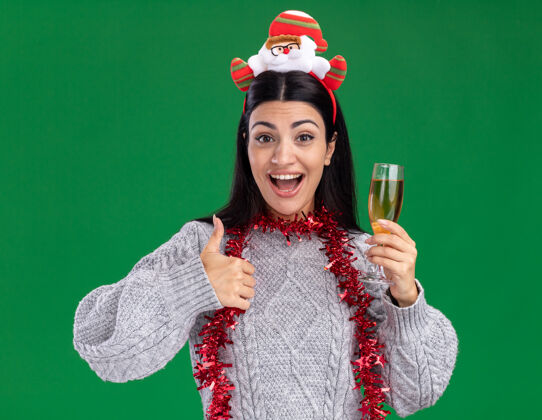 显示年轻的白人女孩戴着圣诞老人的头带 脖子上戴着金属丝的花环 手里拿着一杯香槟 在绿色的墙上孤立地竖起大拇指香槟圣诞老人脖子