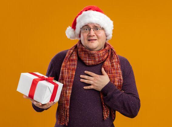 橙色兴奋的成年男子戴着眼镜 戴着圣诞帽 脖子上围着围巾 手里拿着礼包 看着镜头做着感谢的手势 孤立在橙色背景上围巾做围着