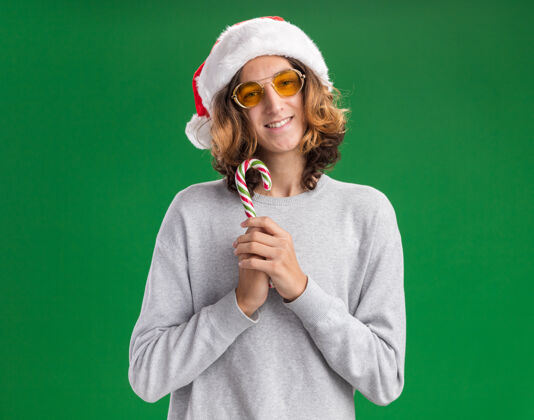 抱着快乐的年轻人戴着圣诞老人的帽子 戴着黄色的眼镜 拿着糖果手杖 站在绿色的背景下 面带微笑地看着相机圣诞节糖果眼镜