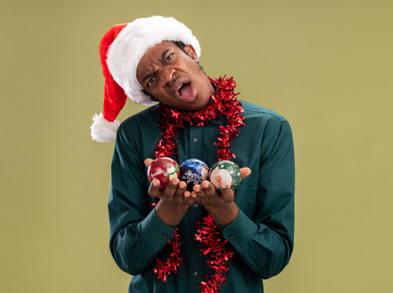 站着不满意的非洲裔美国人戴着圣诞帽 戴着花环 拿着圣诞球 站在绿色背景下 表情困惑地看着摄像机圣诞老人不满意帽子
