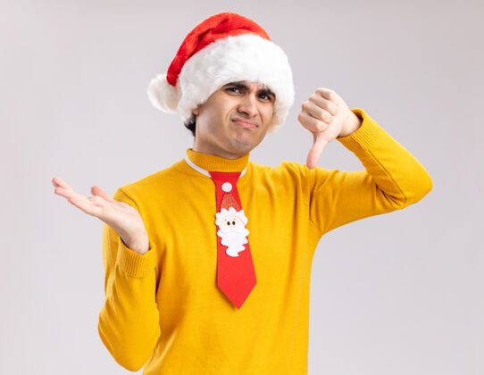 介绍一个穿着黄色高领毛衣 戴着圣诞老人帽 打着滑稽领带的年轻人看着摄像机 用手举着大拇指 站在白色背景上不高兴地展示着什么东西向下圣诞老人拇指