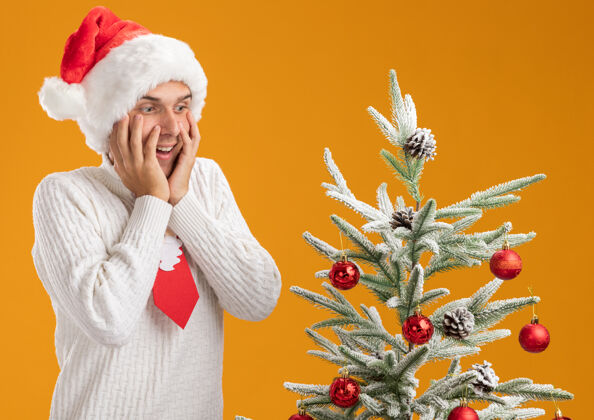 年轻的年轻帅哥戴着圣诞帽 打着圣诞老人的领带 站在装饰好的圣诞树旁 手放在脸上 孤零零地看着橙色的墙上靠近领带印象深刻
