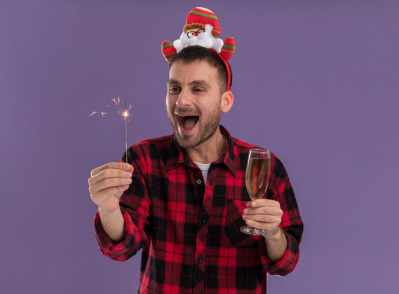 圣诞老人印象深刻的年轻白人男子戴着圣诞老人的头带举行假日火花和香槟杯看着火花孤立在紫色墙上复制空间圣诞老人香槟圣诞节
