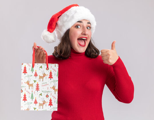 帽子快乐的年轻女孩穿着红色毛衣 戴着圣诞帽 手里拿着彩色纸袋和圣诞礼物 面带微笑地看着相机 竖起大拇指站在白色背景上毛衣站着年轻人