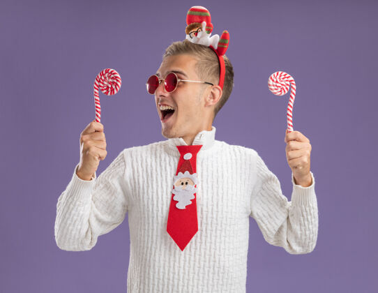 得到年轻帅哥戴着圣诞老人的头带和领带 戴着眼镜 拿着圣诞糖果手杖 看着其中一个正准备吃掉它的紫色背景帅哥看眼镜