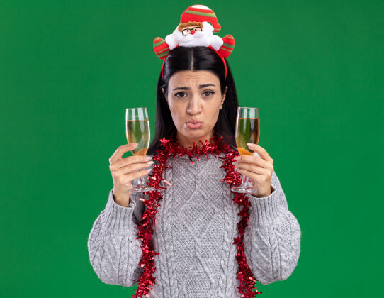 圣诞老人困惑的年轻白人女孩戴着圣诞老人的头带 脖子上戴着金箔花环 手里拿着两杯香槟 隔离在绿色的墙上圣诞节脖子年轻