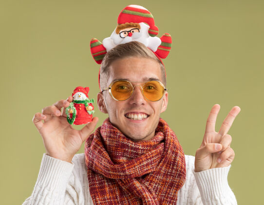 围巾快乐的年轻帅哥戴着圣诞老人的头带和围巾拿着雪人的圣诞饰品做着和平的标志孤立在橄榄绿的墙上雪人头带圣诞老人