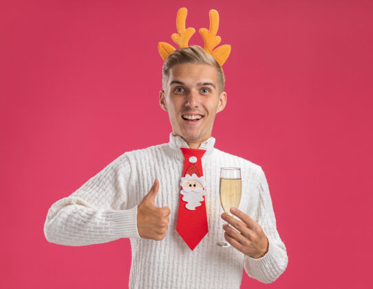 圣诞老人快乐的年轻帅哥戴着驯鹿的鹿角头带 打着圣诞老人的领带 手里拿着一杯香槟 大拇指竖起 孤零零地站在粉红色的墙上玻璃帅哥拇指