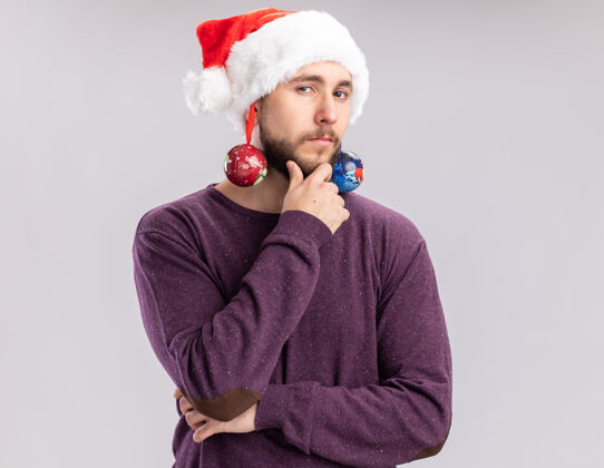 帽子穿着紫色毛衣 戴着圣诞帽的年轻人戴着滑稽的眼镜 耳朵上戴着圣诞球 带着沉思的表情看着镜头 站在白色的背景下思考站着沉思搞笑