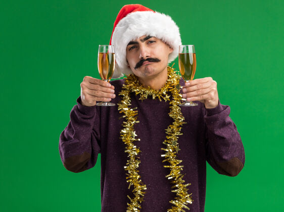 金属片年轻的胡子男人戴着圣诞老人的帽子 脖子上戴着金属丝 手里拿着两杯香槟 站在绿色的背景下自信地看着镜头圣诞老人眼镜站着