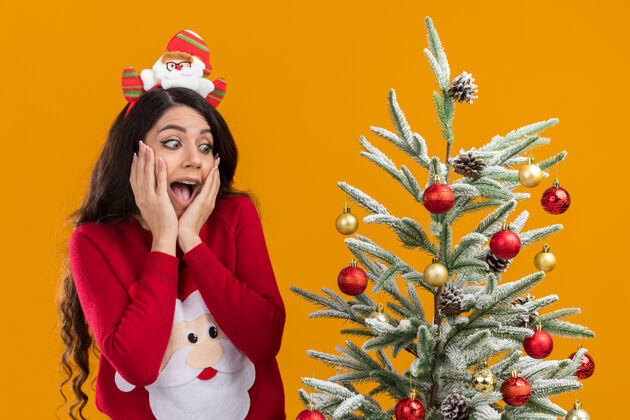 手兴奋的年轻漂亮女孩戴着圣诞老人的头带和毛衣站在装饰过的圣诞树旁 手放在脸上看着橙色背景下孤立的一面附近装饰快乐
