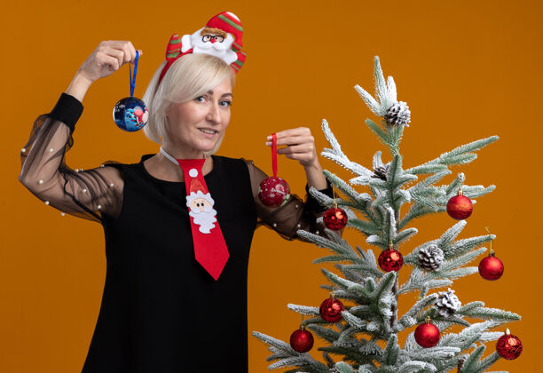 站着微笑着的中年金发女人戴着圣诞老人的头带和领带站在装饰过的圣诞树旁 手里拿着圣诞饰品 看着橙色背景上孤立的摄像头相机装饰圣诞树
