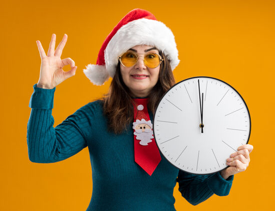 帽子戴着太阳眼镜 戴着圣诞帽 系着圣诞领带 拿着时钟 打着ok手势的成年白种女人高兴地站在橙色背景上 留有复制空间好的成人圣诞老人
