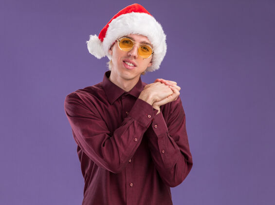 金发戴着圣诞帽戴着眼镜的年轻金发男人高兴地看着相机 手在紫色背景上与世隔绝 留着复印空间保持手年轻