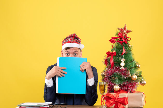 前面商务人士的正面图用文件夹遮住他的脸 放在圣诞树旁的桌子上 黄色的墙上摆着礼物文件桌子人