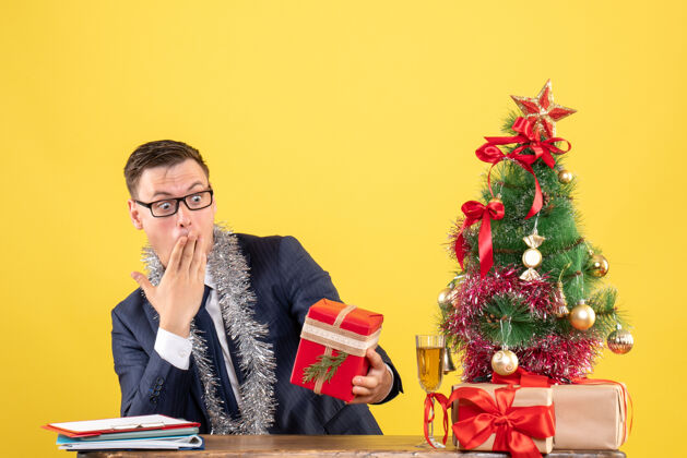 礼物令人惊讶的男人坐在圣诞树旁的桌子前看着他的礼物 黄色的墙上挂着礼物惊奇快乐圣诞节