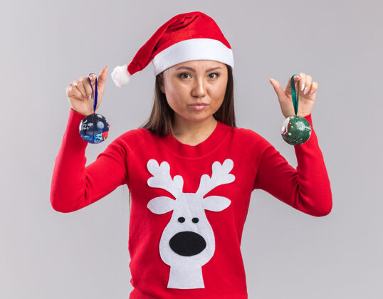 树看着镜头年轻的亚洲女孩戴着圣诞帽拿着毛衣拿着圣诞树球孤立在白色背景上女孩年轻球