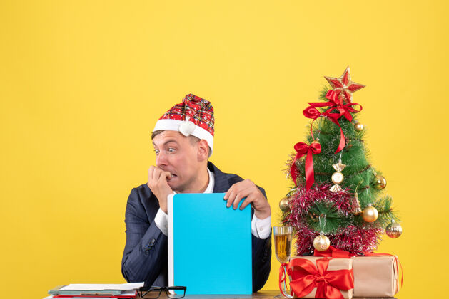 圣诞节焦躁不安的商人坐在圣诞树旁的桌子前 黄色的墙上摆着礼物礼物桌子人