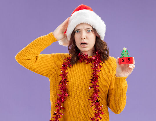 头焦虑的斯拉夫女孩戴着圣诞帽 脖子上戴着花环 手放在头上 手拿着圣诞树饰物 背景是紫色的 有复制空间装饰品周围手