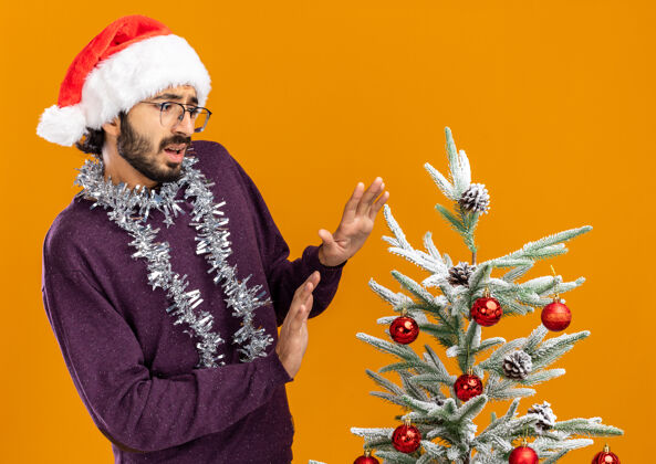 抱着站在圣诞树旁的年轻帅哥 戴着圣诞帽 脖子上戴着花环 站在橙色背景下 手伸向镜头站着树帽子