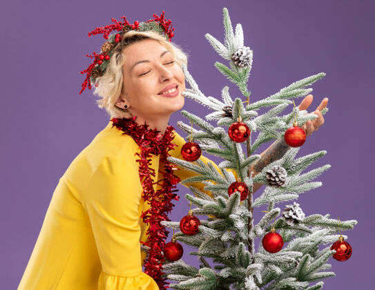 视图微笑的金发女郎头戴圣诞花环 脖子上戴着金属丝花环 站在装饰过的圣诞树旁的侧视图中 闭着眼睛拥抱着它 紫色背景上是孤立的头镀金侧面