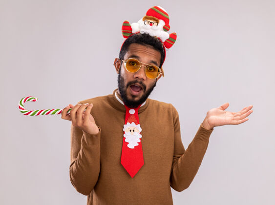 手杖一个非洲裔美国人 穿着棕色毛衣 头上戴着圣诞老人的戒指 打着有趣的红色领带 手里拿着糖果手杖 困惑地看着摄像机 举起双臂站在白色背景上非洲抱着糖果