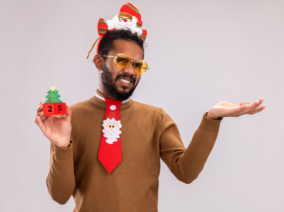 头一个非洲裔美国人 穿着棕色毛衣 头戴圣诞老人圈 打着有趣的红色领带 手里拿着玩具方块 日期是25岁 他看着相机 不高兴地伸出胳膊站在白色背景上日期举行毛衣