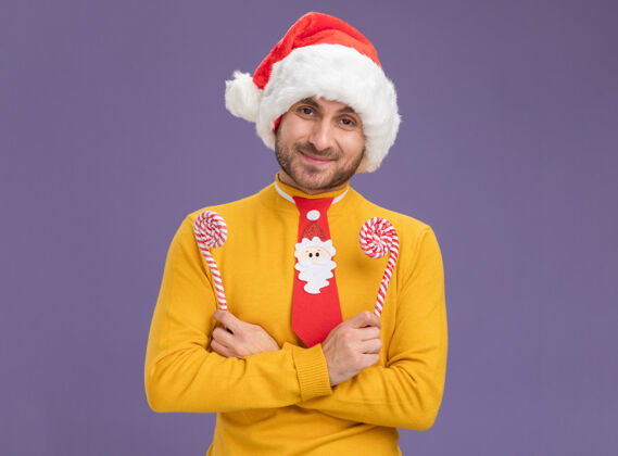 看高兴的年轻白人男子戴着圣诞帽 打着领带 双手交叉 手持圣诞糖果手杖 看着隔离在紫色背景上的相机 还有复印空间帽子手杖年轻