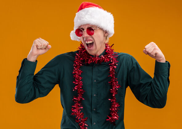 眼镜快乐的年轻金发男子戴着圣诞帽 戴着眼镜 脖子上戴着金箔花环 做着“是”的手势 闭着眼睛 孤立地站在橙色背景上脖子圣诞老人年轻