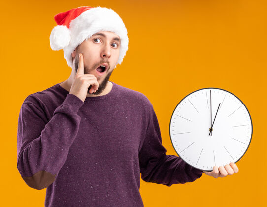 站着一个穿着紫色毛衣 戴着圣诞帽 拿着挂钟的年轻人站在橙色的背景上 看上去既惊讶又惊讶惊讶帽子毛衣