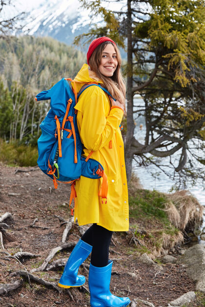 风景很高兴背着背包的年轻女模特戴着红色的头饰 黄色的雨衣和蓝橡胶靴享受杉木徒步旅行