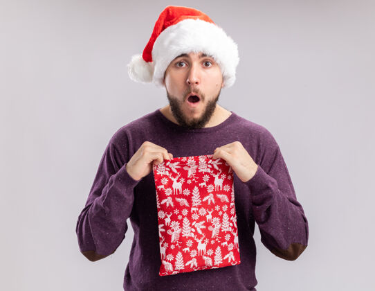 圣诞老人快乐的年轻人穿着紫色毛衣 戴着圣诞老人的帽子 手里拿着红包 手里拿着礼物 站在白色的背景上 惊讶地看着镜头惊讶年轻人礼物