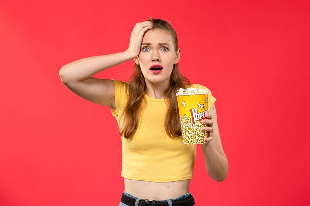 色彩正面图年轻的女性在电影院拿着美味的爆米花在红墙电影院女颜色女性摆姿势爆米花