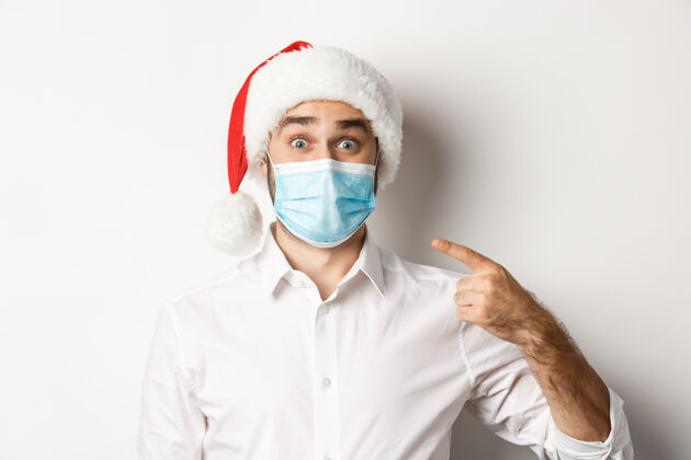 面具冠状病毒的概念 检疫和寒假兴奋的家伙在圣诞帽指着他的面具 站着医疗西装商人