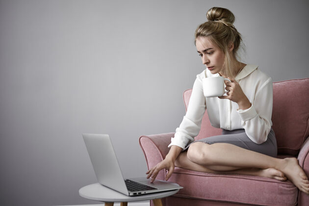 肖像现代科技 工作和休闲的概念严肃的年轻女商人在早上喝咖啡 上班前查看电子邮件 坐在扶手椅上打开笔记本电脑 拿着大杯子杯子人舒适