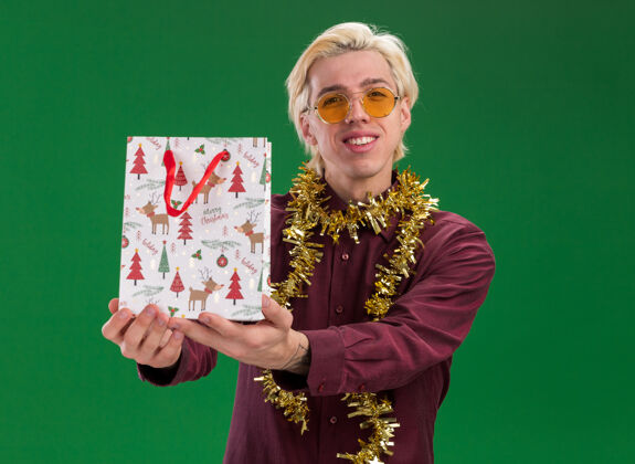 微笑微笑的年轻金发男子戴着眼镜 脖子上戴着金属丝花环 向镜头伸出圣诞礼包 看着隔离在绿色背景上的相机包伸展圣诞节