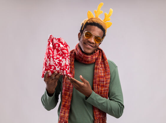 举行一个非洲裔美国人 戴着滑稽的边 戴着鹿角 脖子上围着围巾 手里拿着红色的圣诞老人包 手里拿着礼物 看着相机微笑着用食指指着它站在白色的背景上周围索引圣诞老人