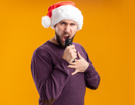 兴奋穿着紫色毛衣 戴着圣诞帽的年轻人站在橙色背景下对着麦克风唱歌 兴奋而快乐帽子麦克风站着