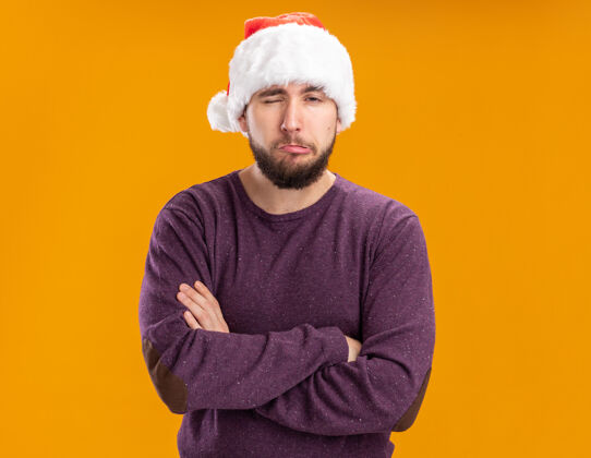 圣诞老人一个穿着紫色毛衣 戴着圣诞帽的年轻人站在橙色的背景下 不高兴地歪着嘴 带着失望的表情看着摄像机制造嘴巴不高兴