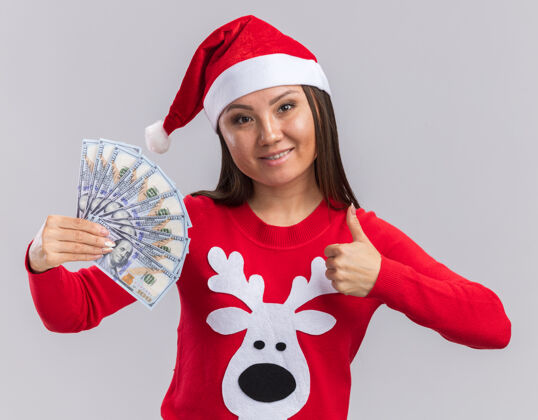 圣诞节笑容满面的年轻亚洲女孩戴着圣诞帽 穿着毛衣 手里拿着现金 在白色背景上孤立地竖起大拇指女孩现金拇指