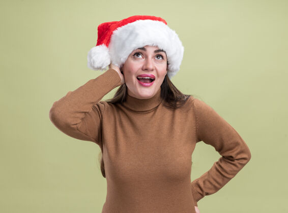 帽子高兴地看着年轻漂亮的女孩戴着圣诞帽把双手放在脸颊和臀部隔离在橄榄绿的背景脸颊放高兴