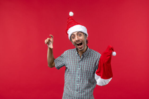 快乐红墙上举着红色圣诞帽的年轻人的正视图成人人脸