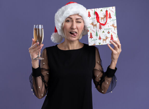 看自信的中年金发女人戴着圣诞帽看着相机拿着一杯香槟摸着头拿着圣诞礼包眨眼 在紫色背景上露出孤立的舌头拿着香槟舌头