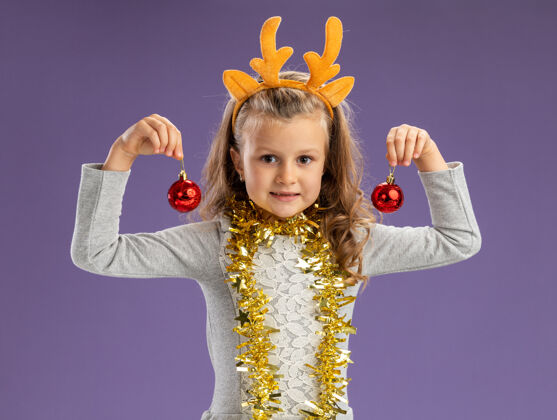 女孩高兴的小女孩戴着圣诞发箍 脖子上戴着花环 手里拿着蓝色背景上孤立的圣诞球小圣诞快乐脖子