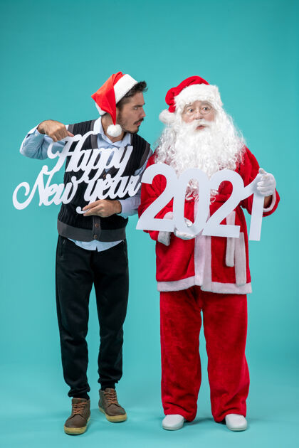 男性圣诞老人的正面图 男性手持新年快乐和蓝色墙上的购物袋圣诞老人帽子圣诞老人
