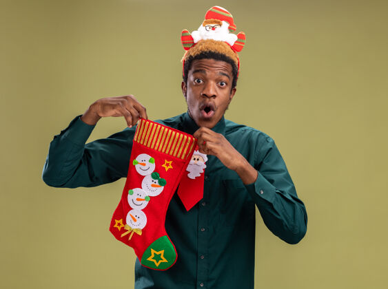 惊讶一个非洲裔美国人 戴着有趣的圣诞老人戒指 打着红色领带 手里拿着圣诞长袜 站在绿色的背景上 惊讶地看着镜头圣诞老人圣诞节美国