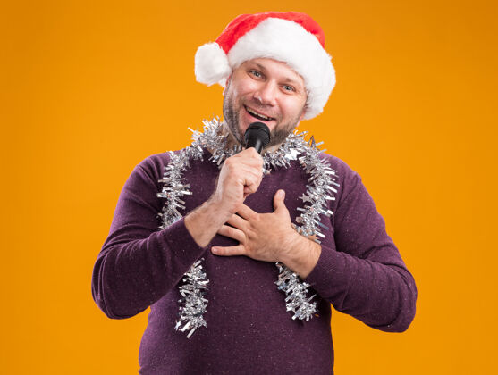 花环快乐的中年男子戴着圣诞帽 脖子上戴着金箔花环 对着麦克风说话 看着相机 手放在胸前 在橙色背景下隔离开来麦克风谈话胸部
