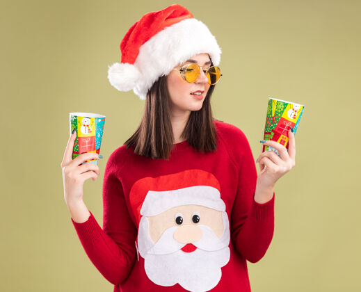 高兴高兴的年轻漂亮的白人女孩穿着圣诞老人毛衣和帽子戴着眼镜拿着塑料圣诞杯看着其中一个孤立的橄榄绿背景女孩塑料举行